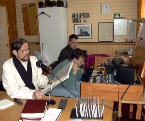 Pappi lukee pöydän ääressä mikrofoniin, taustalla äänittäjät