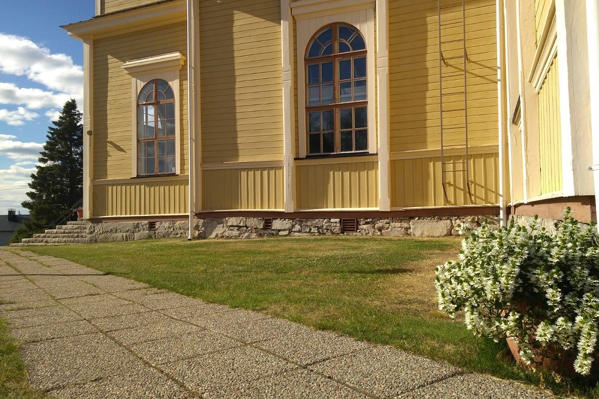 keltainen puukirkko, edessä nurmikkoa ja laatoitettu käytävä