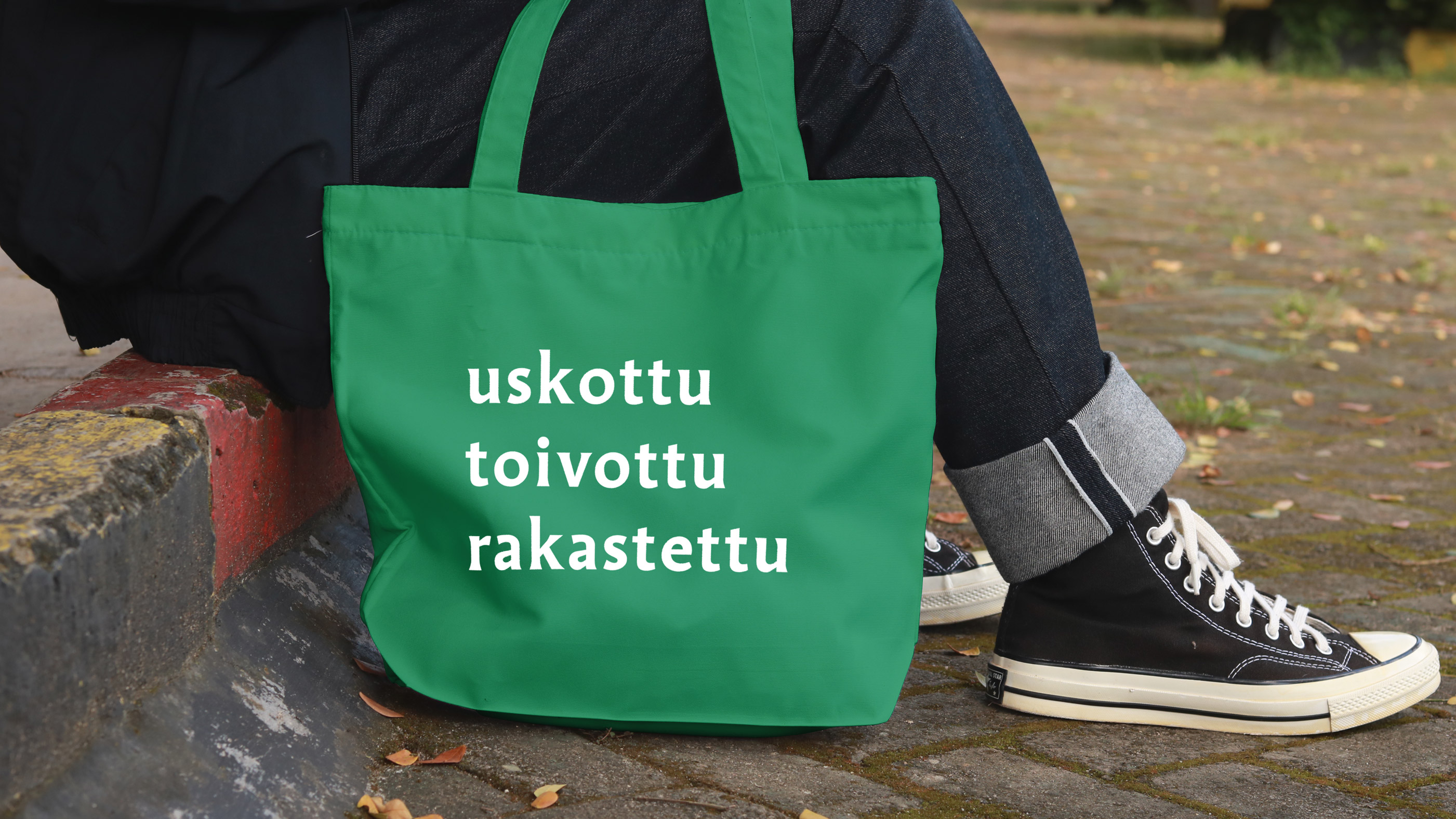 Ihminen istuu jalkakäytävän reunalla, vieressä kassi jossa teksti uskottu toivottu rakastettu
